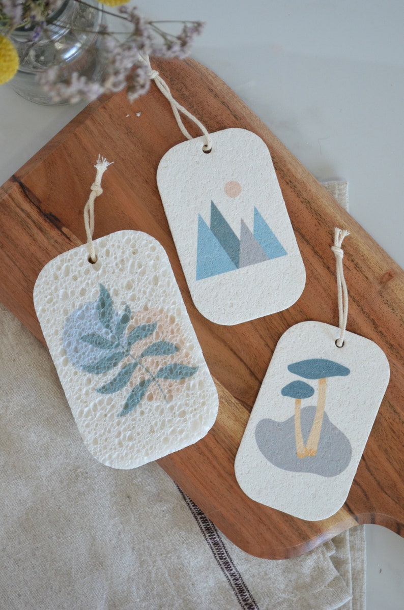 May include: Three white, rectangular, textured, hanging ornaments with different designs. One ornament features a blue and green leaf, another features a blue and gray mountain range, and the last features a gray mushroom with a yellow stem.