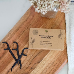 May include: A rectangular cardboard box labeled Charcoal Dental Floss Picks with 50 count, featuring black text and botanical illustrations. Two black plastic dental floss picks with textured handles are positioned next to the box.