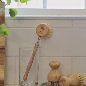 May include: Three wooden dish brushes with natural bristles. The brushes are on a light green countertop. The largest brush has a wooden handle and a round wooden head with the text "ME Mother Earth" printed on it.