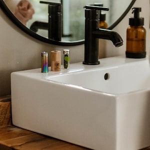 May include: A white porcelain bathroom sink with a black faucet and three small bottles on the counter. The bottles are different colors and have different designs.