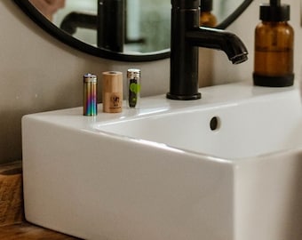 May include: A white porcelain bathroom sink with a black faucet and three small bottles on the counter. The bottles are different colors and have different designs.