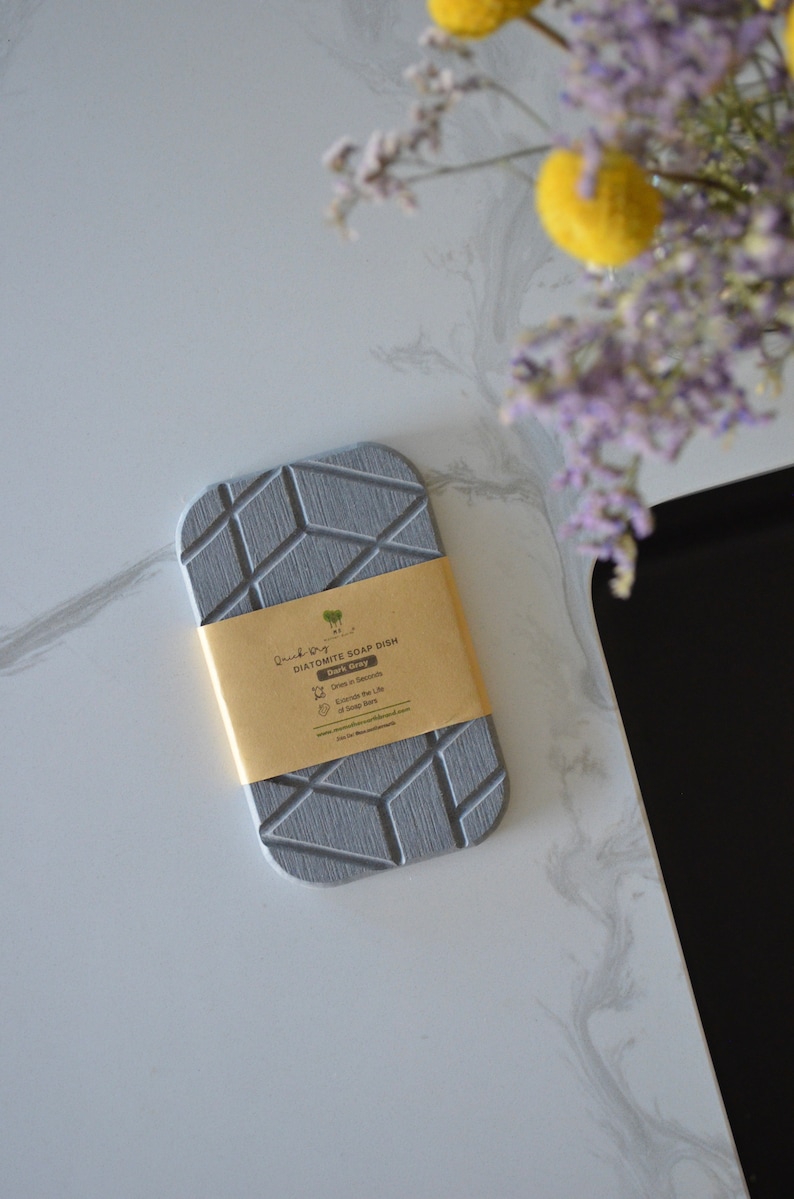 K&ouml;nnte beinhalten: Eine graue Diatomeenerde-Seifenschale mit geometrischem Muster. Die Schale ist mit "Diatomite Soap Dish Dark Grey" beschriftet und tr&auml;gt die Aufschrift "Drains in Seconds" und "Extends the Life of Soap Bars".