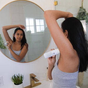 Peut inclure: Une personne s'applique du d&eacute;odorant sous le bras dans une salle de bain. Le d&eacute;odorant est un b&acirc;ton blanc avec du texte noir qui indique "D&eacute;odorant sans aluminium".