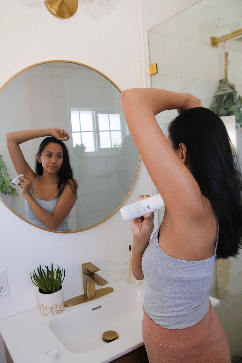 Peut inclure: Une personne s'applique du d&eacute;odorant sous le bras dans une salle de bain. Le d&eacute;odorant est un b&acirc;ton blanc avec du texte noir qui indique "D&eacute;odorant sans aluminium".