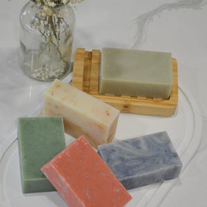 May include: An assortment of handmade soaps in various colors, including green, orange, and blue, arranged on a white tray. A wooden soap dish holds a light green bar. A glass vase with flowers is in the background.