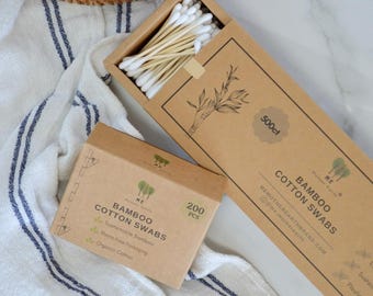 May include: Two rectangular cardboard boxes containing bamboo cotton swabs. The larger open box holds white-tipped swabs, while the smaller closed box is labelled 200 PCS. Both feature the text Bamboo Cotton Swabs and Mother Earth branding.