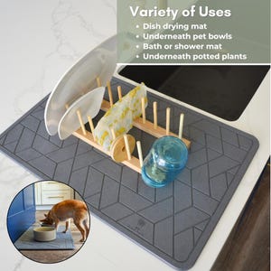 May include: A grey, rectangular, textured silicone mat with a geometric pattern. The mat is placed on a white worktop and has a dish drying rack with dishes and a glass on top of it. A dog is eating from a bowl on a smaller version of the mat in the bottom left corner of the image. The text "Variety of Uses" is in the top right corner of the image with a list of uses for the mat: "Dish drying mat", "Underneath pet bowls", "Bath or shower mat", "Underneath potted plants".