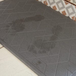May include: A grey, rectangular doormat with a geometric pattern of diamonds. The doormat has a textured surface and the words "The Doormat" are printed in white on the bottom right corner.