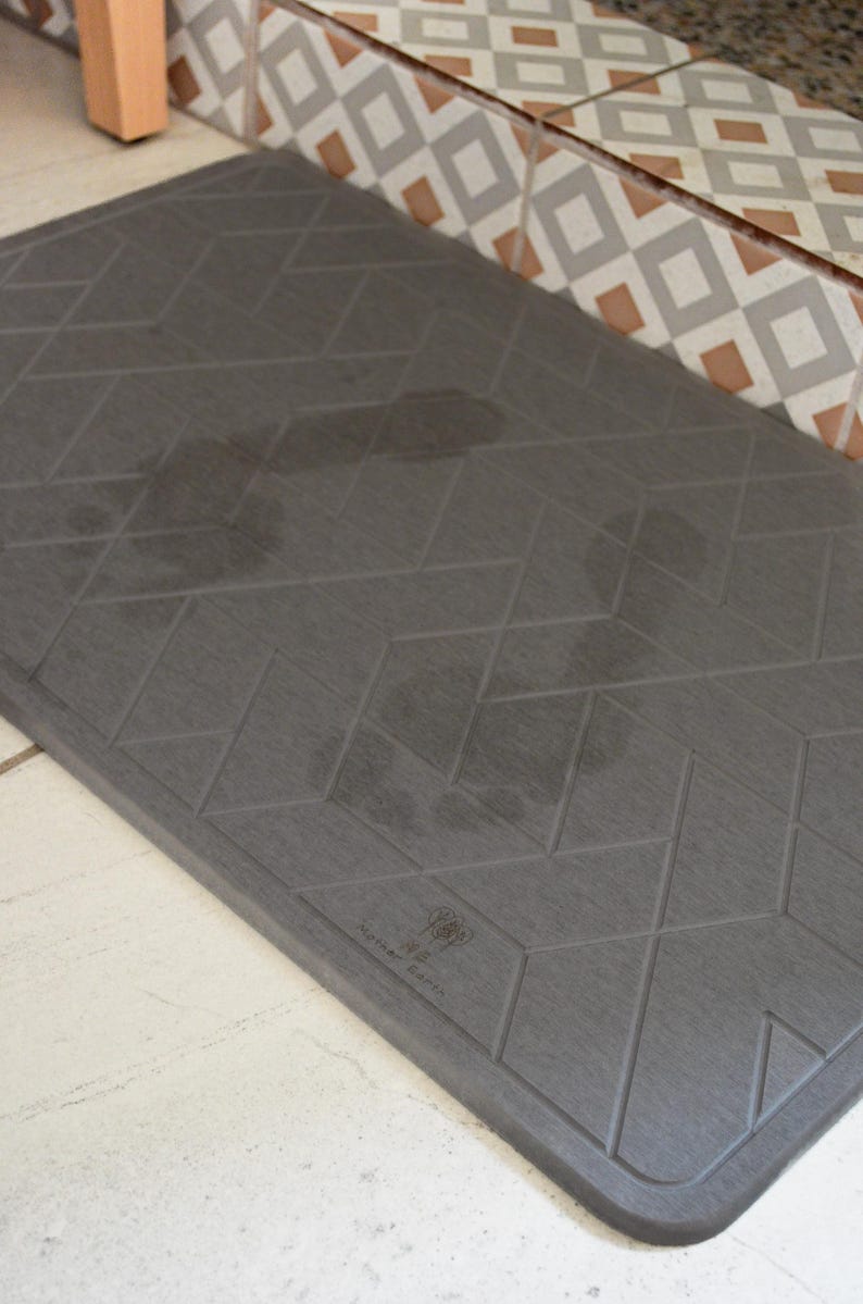 May include: A grey, rectangular doormat with a geometric pattern of diamonds. The doormat has a textured surface and the words "The Doormat" are printed in white on the bottom right corner.