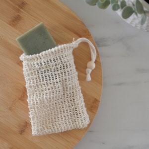 May include: A white mesh soap bag with a wooden bead drawstring closure, containing a green bar of soap, on a wooden cutting board.