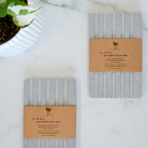 May include: Two gray diatomite soap dishes with brown paper labels. The labels read "Quick Dry Diatomite Soap Dish" and include the company name "HE Mother Earth".