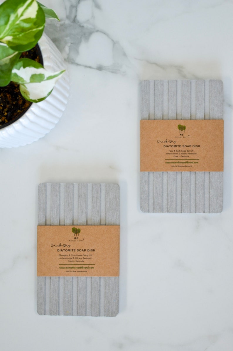 May include: Two gray diatomite soap dishes with brown paper labels. The labels read "Quick Dry Diatomite Soap Dish" and include the company name "HE Mother Earth".