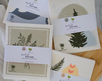 May include: Four sets of three Swedish dish cloths wrapped in white paper bands labeled Mother Earth. The cloths feature various printed designs including a yellow sun, a dark green pine tree, a botanical floral arrangement, and orange moon phases.