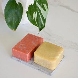 May include: Two bars of soap on a gray soap dish. The soap on the left is pink and labeled "FACE". The soap on the right is yellow and labeled "BODY".