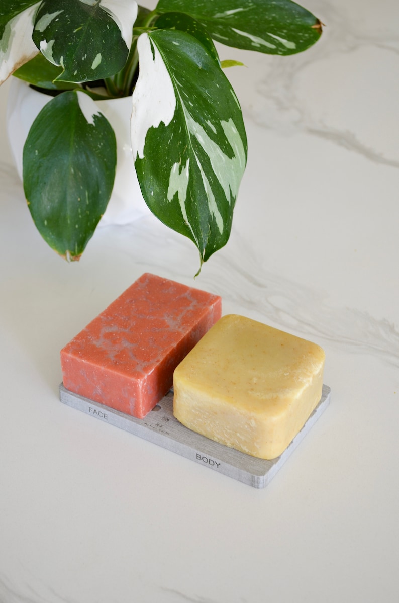May include: Two bars of soap on a gray soap dish. The soap on the left is pink and labeled "FACE". The soap on the right is yellow and labeled "BODY".