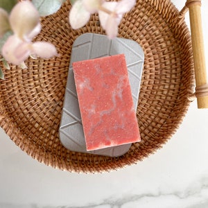 K&ouml;nnte beinhalten: Ein rosafarbener Seifenblock mit gesprenkelter Textur liegt auf einem grauen Seifensch&auml;lchen in einem geflochtenen Weidenkorb.