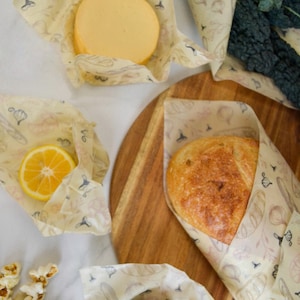 May include: A set of four reusable food wraps with a floral pattern. The wraps are made of beeswax, jojoba oil, and tree resin. The wraps are shown wrapped around a block of cheese, a lemon, a loaf of bread, and an avocado.