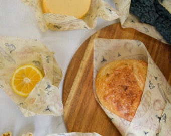 May include: A set of four reusable food wraps with a floral pattern. The wraps are made of beeswax, jojoba oil, and tree resin. The wraps are shown wrapped around a block of cheese, a lemon, a loaf of bread, and an avocado.