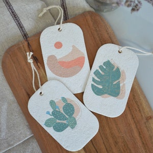 May include: Three rectangular gift tags made of recycled paper with different abstract designs. The tags are white with a light brown speckled texture. The designs are a peach colored abstract shape with a small orange circle, a green monstera leaf with gold veins, and a green cactus with white dots.
