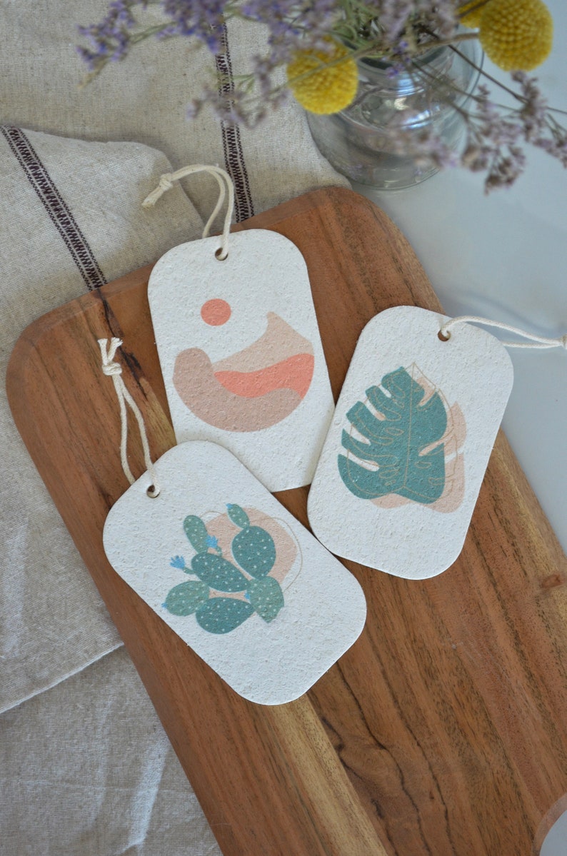 May include: Three rectangular gift tags made of recycled paper with different abstract designs. The tags are white with a light brown speckled texture. The designs are a peach colored abstract shape with a small orange circle, a green monstera leaf with gold veins, and a green cactus with white dots.