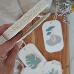 May include: Four ceramic gift tags with different designs. The tags are white with a light brown background and feature a mushroom, a leaf, a cactus, and a geometric shape. The tags are on a wooden surface.