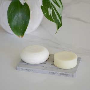 May include: Two white solid shampoo and conditioner bars on a gray soap dish with the words "Shampoo" and "Conditioner" printed on it.