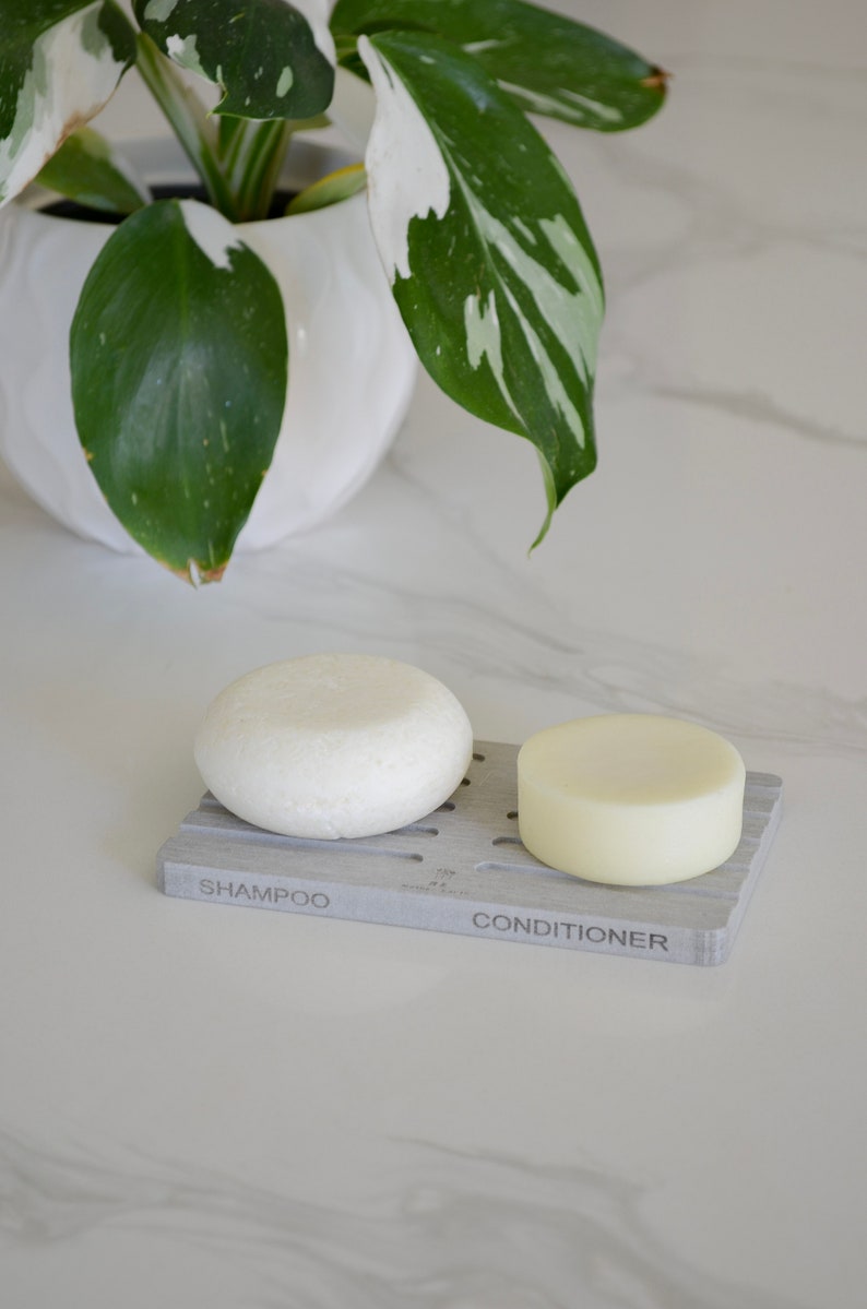 May include: Two white solid shampoo and conditioner bars on a gray soap dish with the words "Shampoo" and "Conditioner" printed on it.