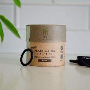 May include: A cylindrical cardboard container of black, plastic-free hair ties. The container has a brown base and a tan lid, with the words "MINI PLASTIC-FREE HAIR TIES" printed on the front. A single black hair tie sits in front of the container.