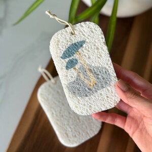 May include: Two white loofah sponges with a string attached. One sponge has a blue and yellow design of a mushroom.