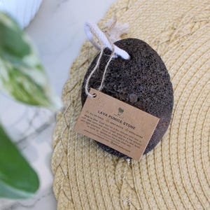 Puede incluir: Una piedra p&oacute;mez de lava de color gris oscuro con una cuerda blanca atada. La piedra est&aacute; sentada sobre una estera de paja tejida. Una etiqueta de papel marr&oacute;n con el texto "Lava Pumice Stone" est&aacute; unida a la piedra.