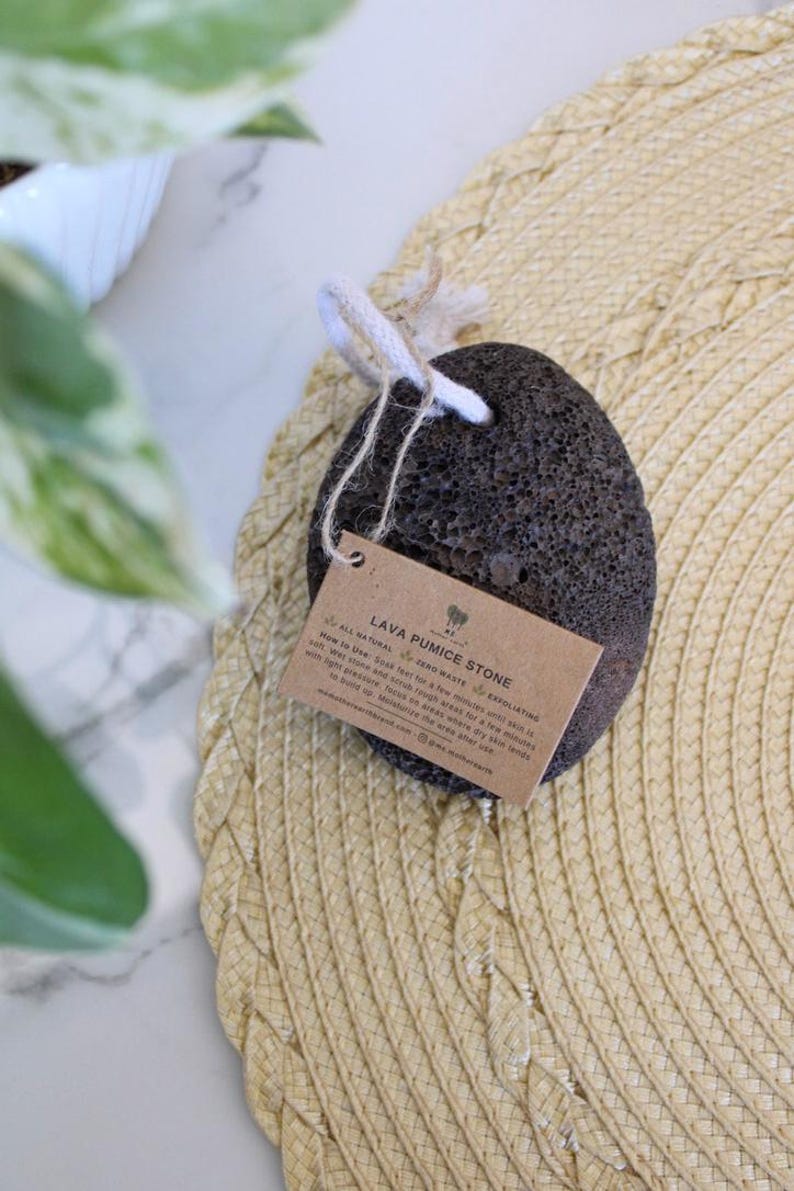 Puede incluir: Una piedra p&oacute;mez de lava de color gris oscuro con una cuerda blanca atada. La piedra est&aacute; sentada sobre una estera de paja tejida. Una etiqueta de papel marr&oacute;n con el texto "Lava Pumice Stone" est&aacute; unida a la piedra.