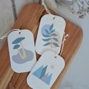 May include: Three rectangular gift tags with string attached. Each tag features a different abstract design in shades of blue, pink, and beige. The designs include mushrooms, a leaf, and mountains.