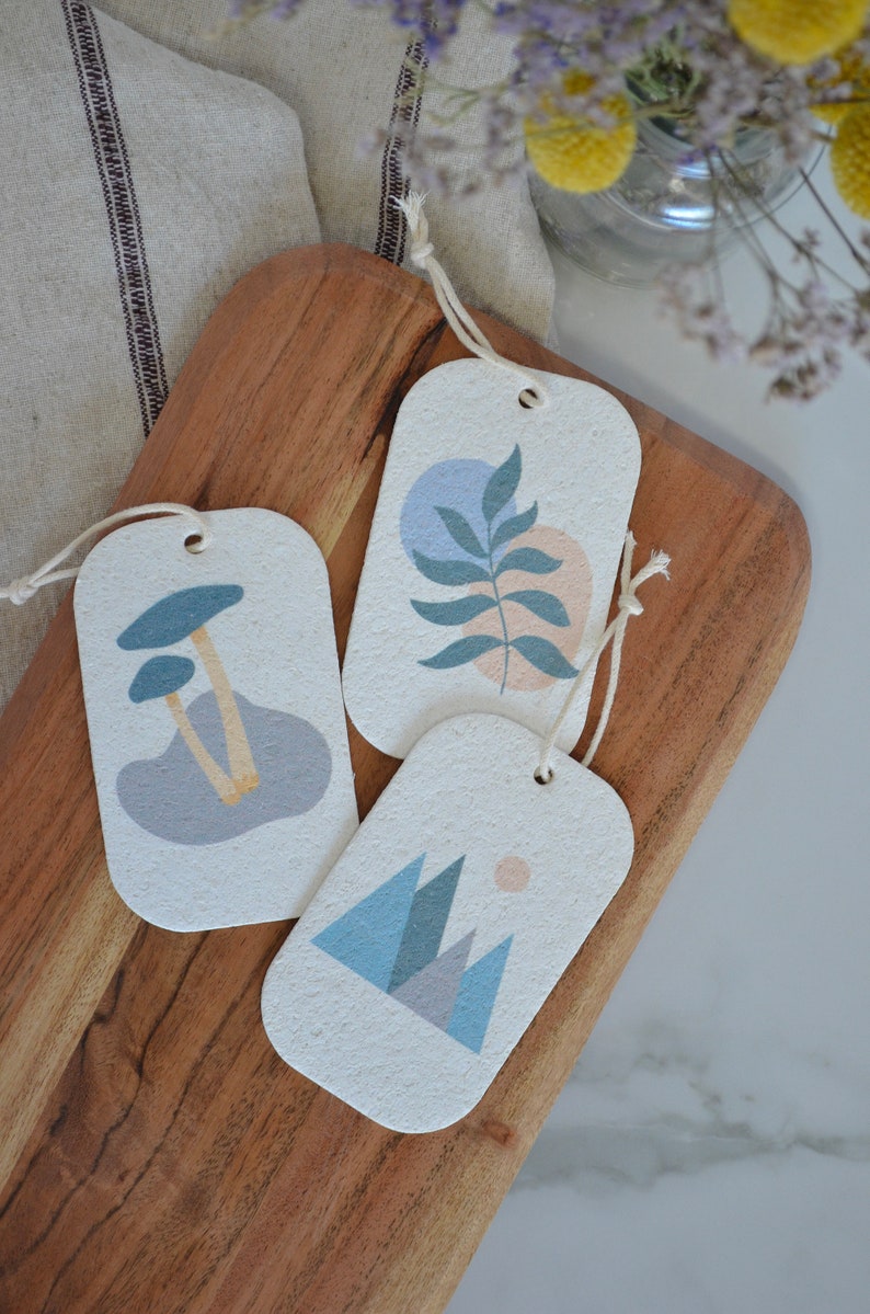 May include: Three rectangular gift tags with string attached. Each tag features a different abstract design in shades of blue, pink, and beige. The designs include mushrooms, a leaf, and mountains.