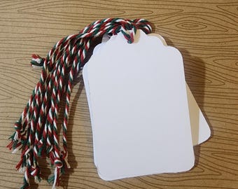 May include: A stack of blank white gift tags with scalloped edges, ready for personalization. They are tied with a festive red, green, and white twisted cord. The tags are on a wood-grain background, perfect for holiday or special occasion use.