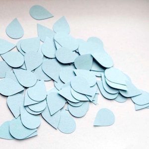 May include: A pile of light blue paper confetti shaped like teardrops. The confetti is scattered on a white surface.