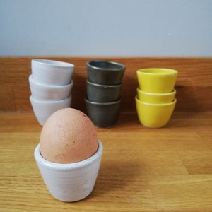 May include: A brown egg sits in a white ceramic egg cup. Stacks of white, grey, and yellow ceramic bowls are behind it. The egg cup and bowls are on a wooden surface, with a grey wall in the background.