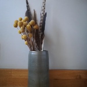 May include: A grey-blue ceramic vase with a tan base, containing an arrangement of yellow globe flowers and feathers. The feathers are in shades of brown and black. The vase is set against a light grey wall and wooden floorboards.