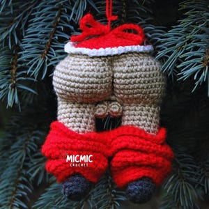 Op de afbeelding: Een handgemaakte kerstversiering in de vorm van een achterwerk, gehaakt in beige, rood en zwart garen. De versiering heeft een rode kerstmuts, een rode broek en zwarte voeten. Het hangt aan een rood touwtje tegen een achtergrond van groene dennennaalden.