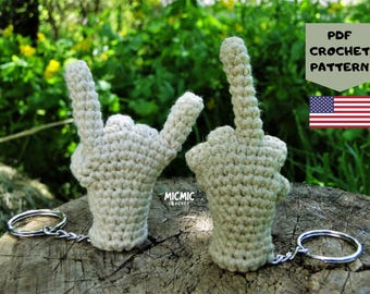 May include: Two crocheted hand keychains, one with the fingers in a rock-and-roll gesture and the other with the middle finger raised. The keychains are beige and attached to silver keyrings. The background includes a green leafy area and a wooden surface.