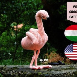 May include: A handcrafted, pink crochet flamingo with a black beak and closed eyes. The flamingo is positioned on a stone surface against a blurred green backdrop. The image includes the text "PDF CROCHET PATTERN".