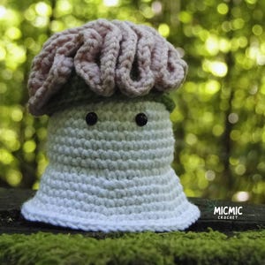 May include: A handmade crochet mushroom figure with a light pink cap and a white and light green body. The mushroom has two black button eyes and is sitting on a mossy surface. The word "MICMIC CROCHET" is visible.