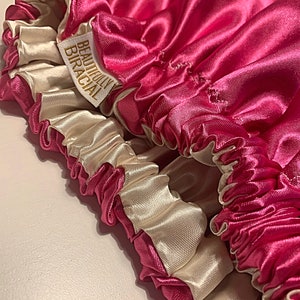 May include: A pink satin hair bonnet with a white satin lining. The bonnet has a gold label that reads "Beautifully Biracial".