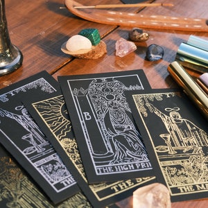 Puede incluir: Cuatro cartas de tarot negras con detalles en pan de oro y plata sobre una mesa de madera. Las cartas son "El Mago", "La Sacerdotisa", "El Sol" y "Los Enamorados".