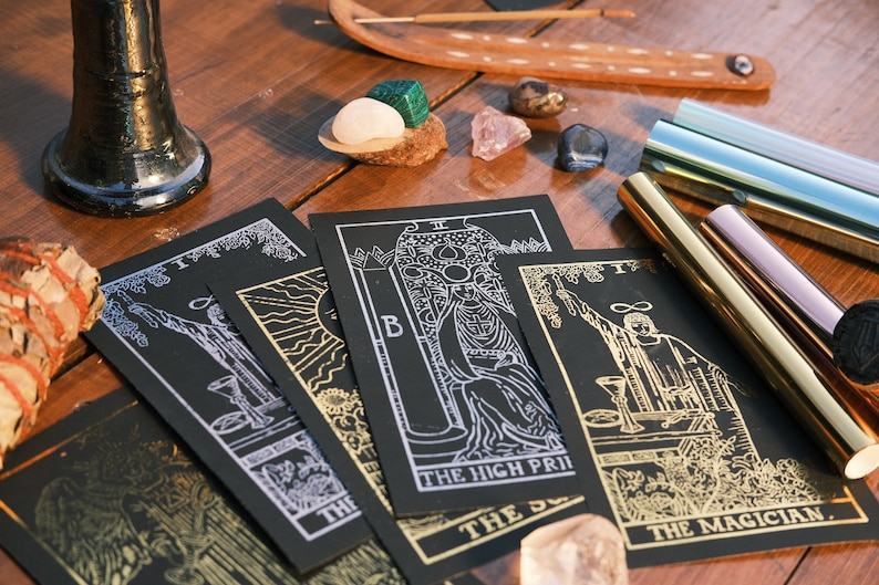 Puede incluir: Cuatro cartas de tarot negras con detalles en pan de oro y plata sobre una mesa de madera. Las cartas son "El Mago", "La Sacerdotisa", "El Sol" y "Los Enamorados".