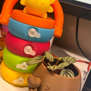 May include: A colorful stacking toy with a yellow sun-shaped top and a curved orange handle. The toy has rings in orange, blue, pink, green, and yellow. A brown planter with a smiling face and heart-shaped arms holds a small plant.