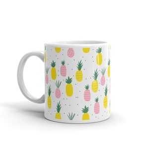 Puede incluir: Taza de cerámica blanca con un patrón repetido de piñas amarillas y rosas sobre un fondo blanco. Las piñas tienen hojas verdes y están rodeadas de pequeñas hojas verdes y puntos blancos.
