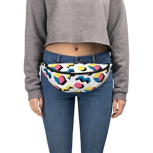 May include: A white fanny pack with a geometric pattern in pink, blue, yellow, and black. The fanny pack is worn around the waist of a person wearing blue jeans.