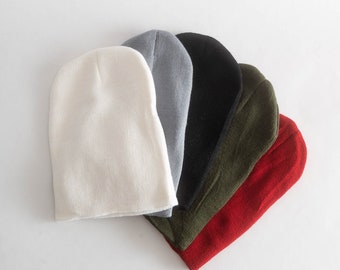 May include: A set of five knit beanies arranged in a fan shape, featuring solid colors including white, light gray, black, olive green, and red. Each beanie is constructed from a soft, ribbed knit fabric with a rounded top and simple design.