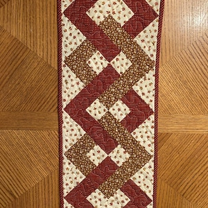 May include: A quilted table runner with a geometric pattern in shades of red, brown, and white. The runner features a repeating diamond pattern with a floral print.