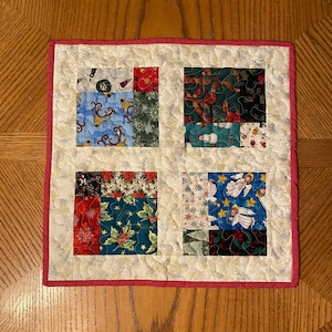 May include: A square quilted Christmas placemat with a red border. The center features four squares with festive patterns including reindeer, holly, angels, and snowflakes. The background is a light cream color.